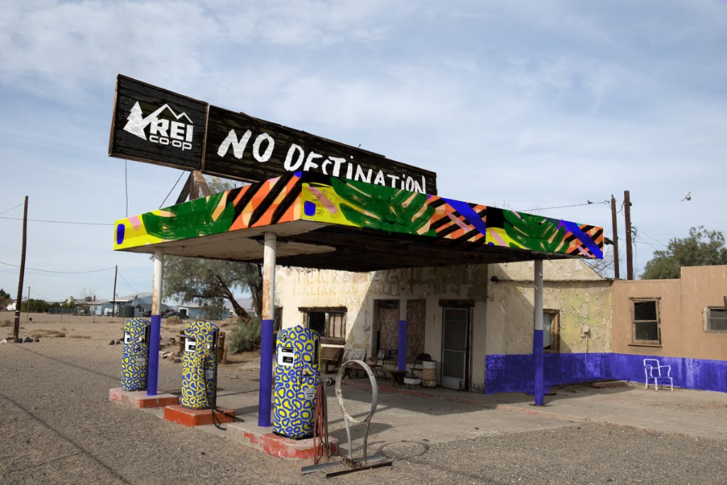 A derelict desert gas station painted over in the No Destination brand — REI Co-op signage and patterned pumps